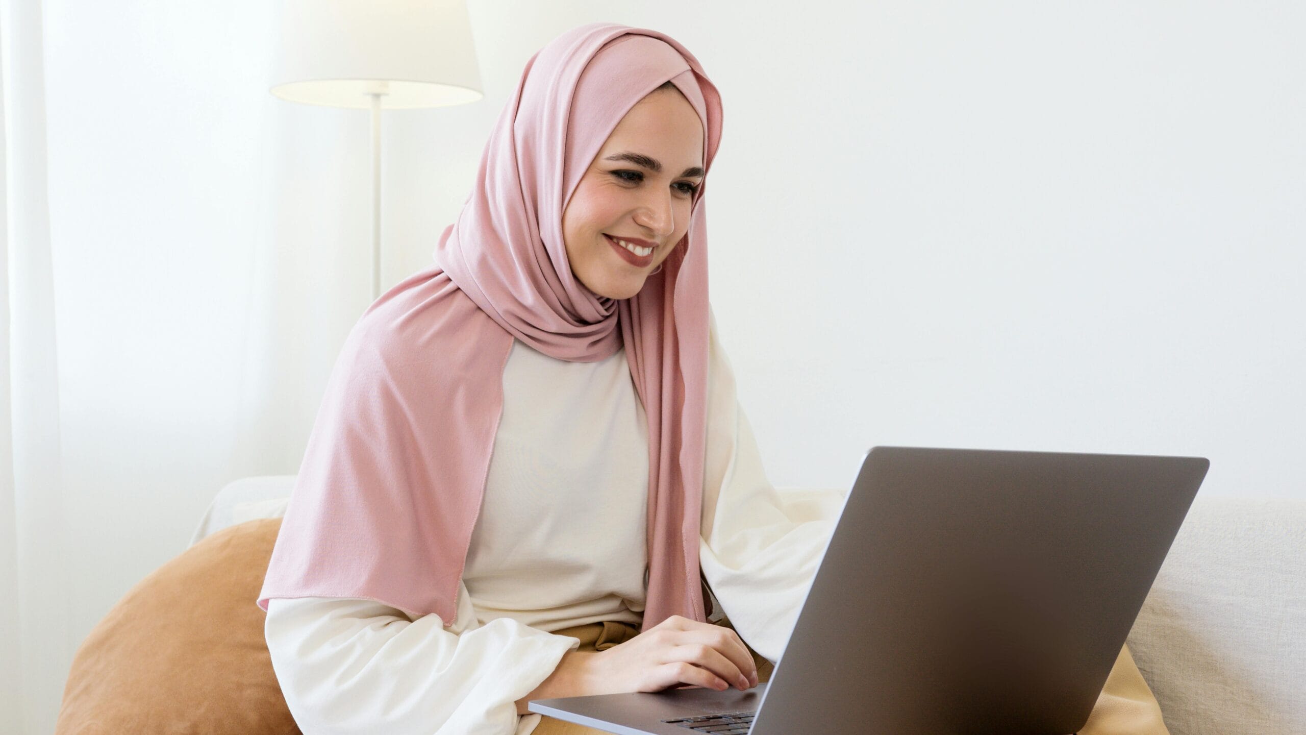 Confident Muslim woman wearing a hijab, smiling while using a laptop in a cozy home setting.